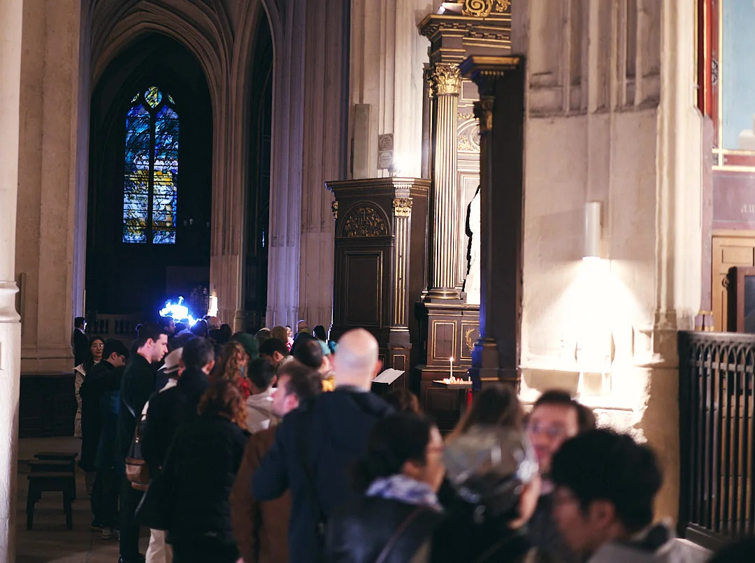 A queue of visitors in the nave of a gothic church, stained glass and gilded woodwork; at the far end, the blue glow of the installation.