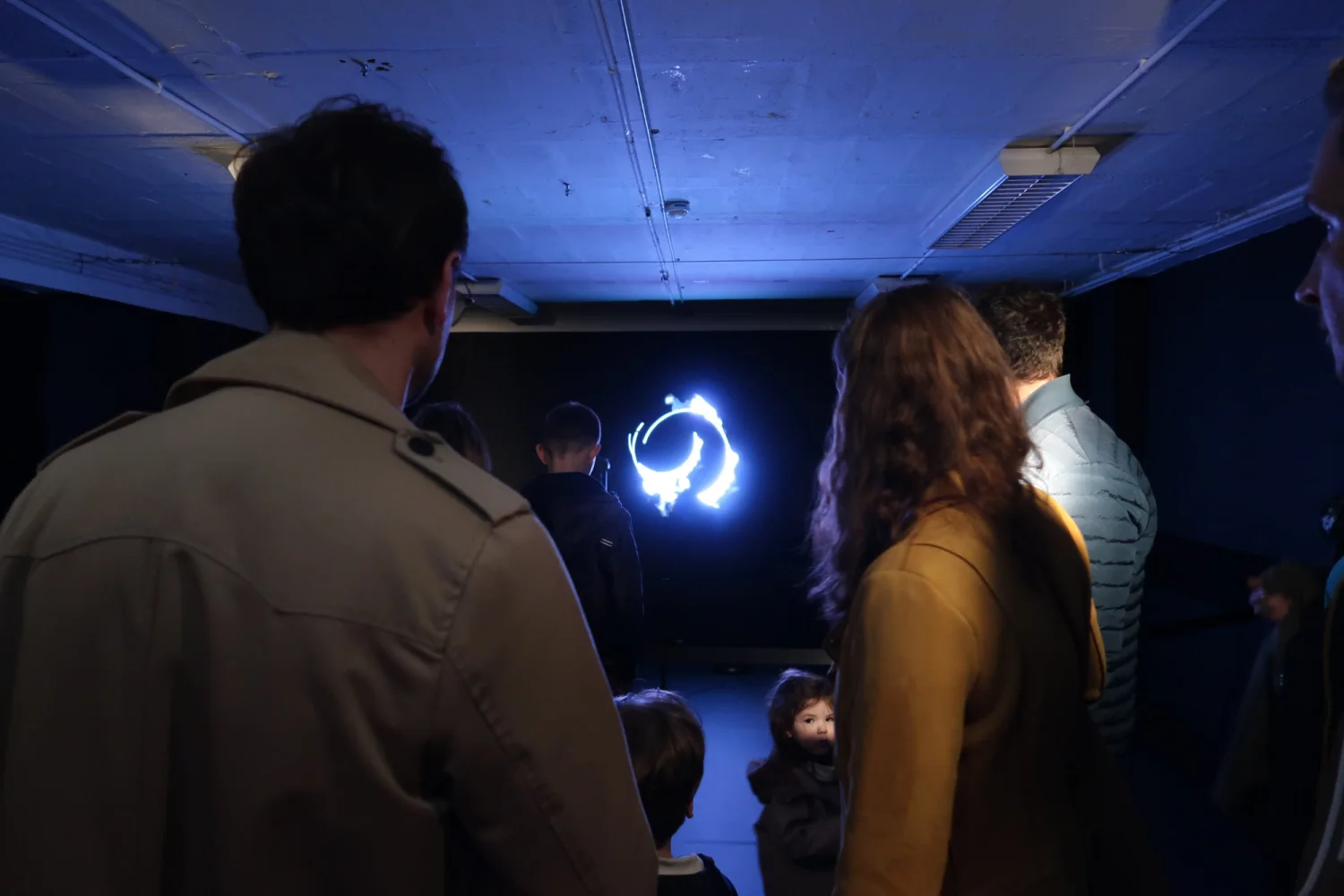 Seen from behind in a darkened room, visitors of all ages watch the blue ring hanging in front of them.