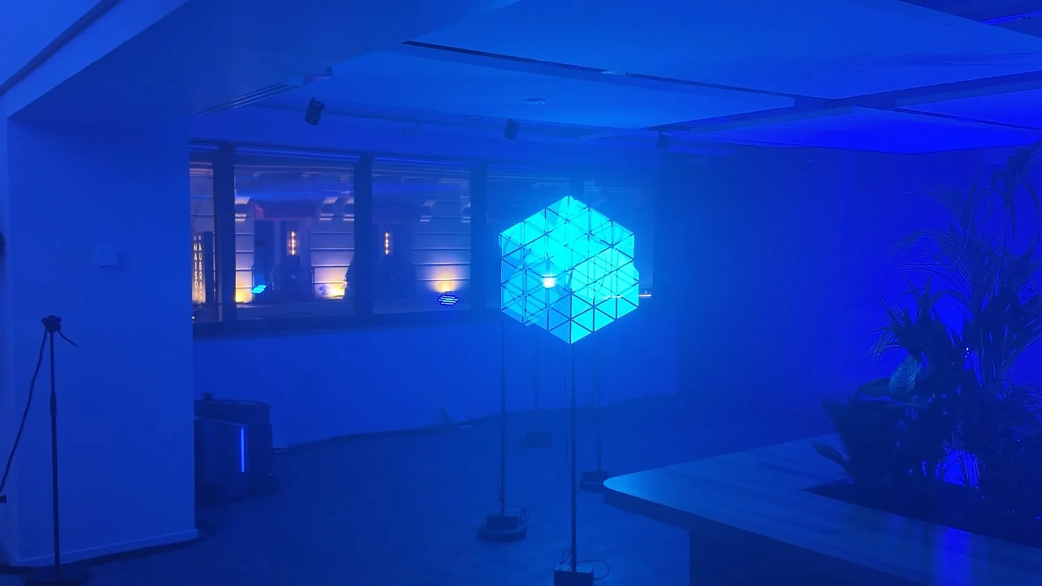 A still from the video: in a room bathed in blue, several propellers on stands recompose a glowing turquoise wireframe cube, suspended in mid-air.