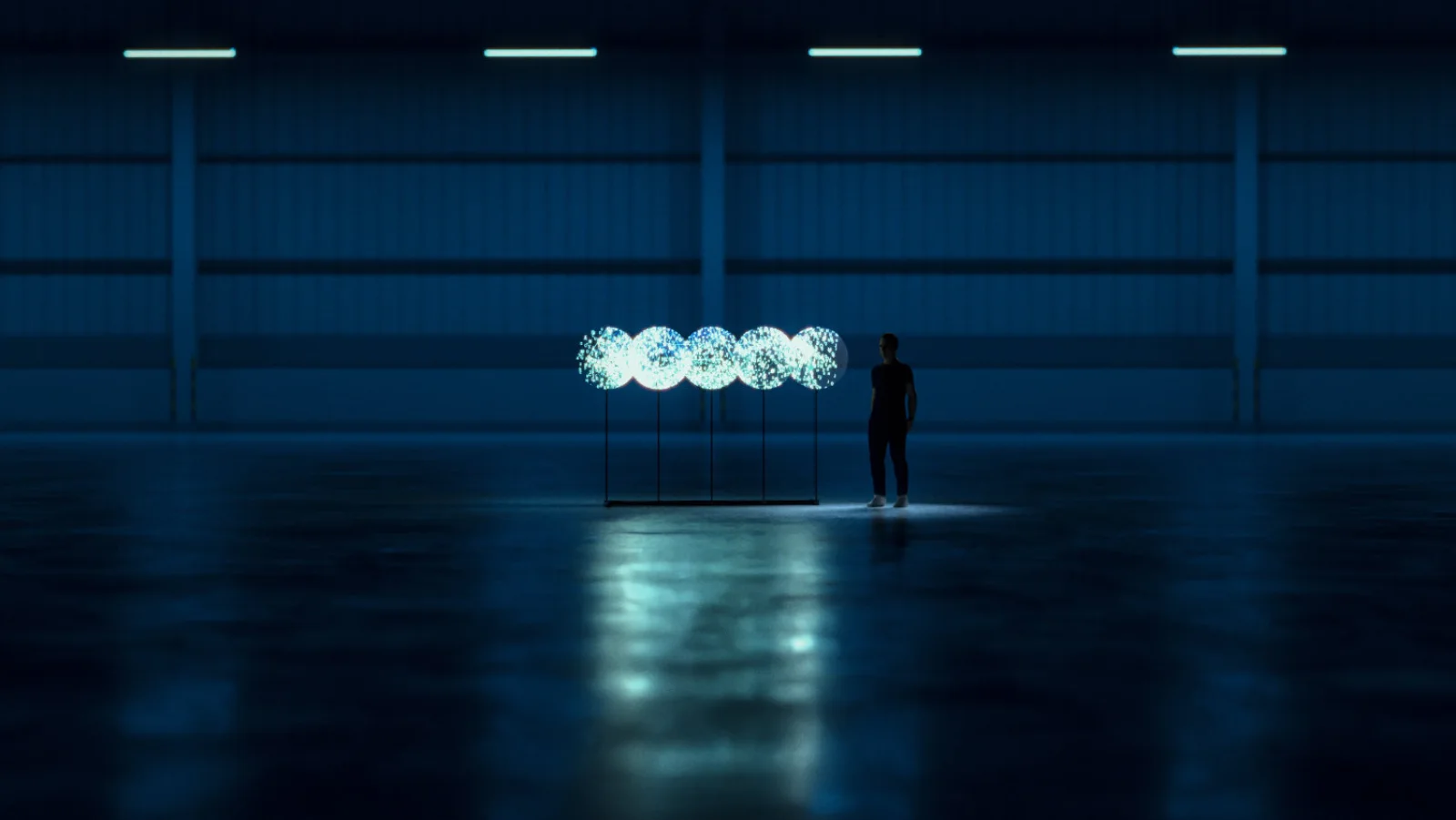 Vue large du hangar : les cinq hélices alignées affichent cinq disques lumineux, une silhouette debout à côté donne l&rsquo;échelle.