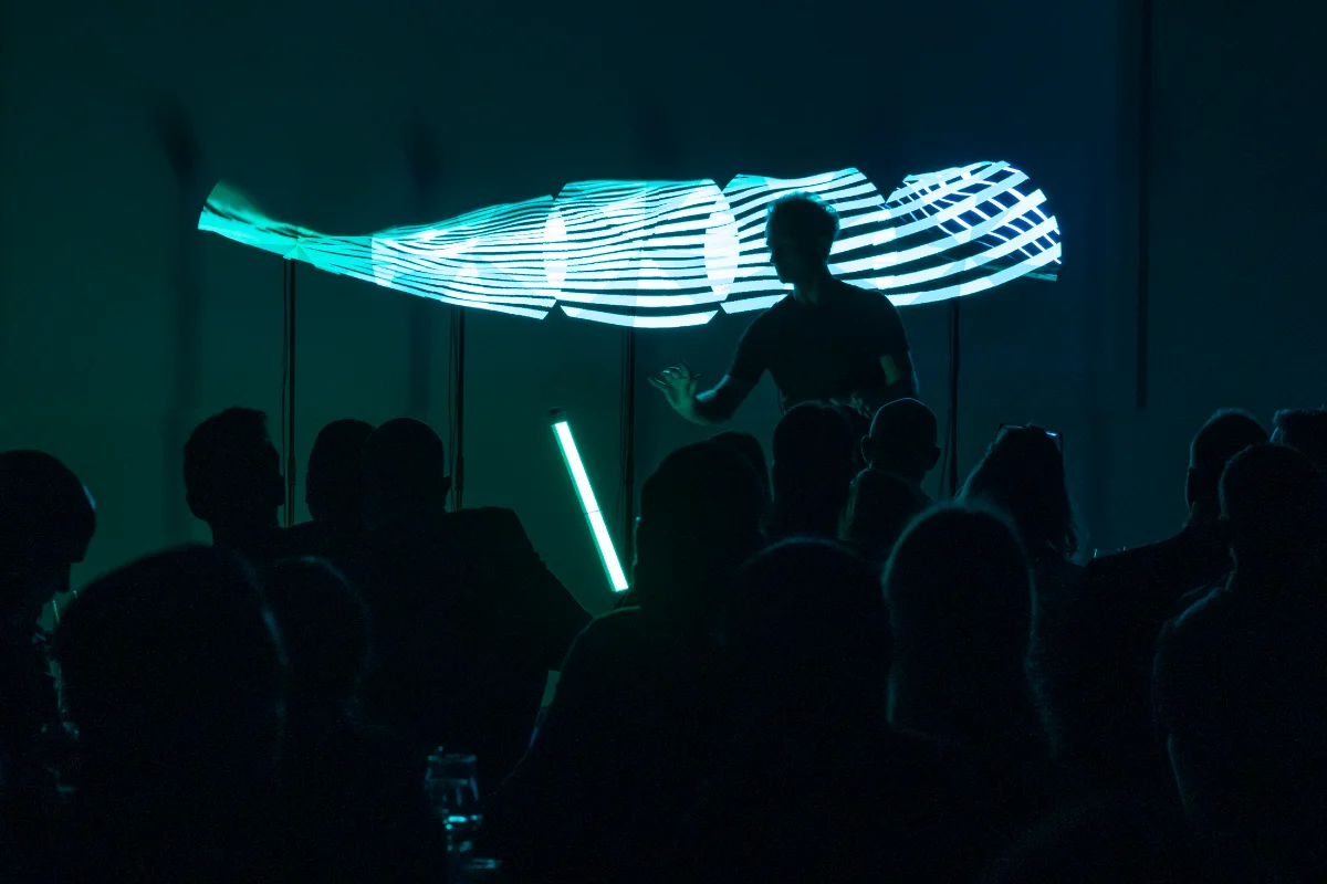 Performance Holo 5 : cinq hélices composent une large image turquoise au-dessus d'un public en silhouette.