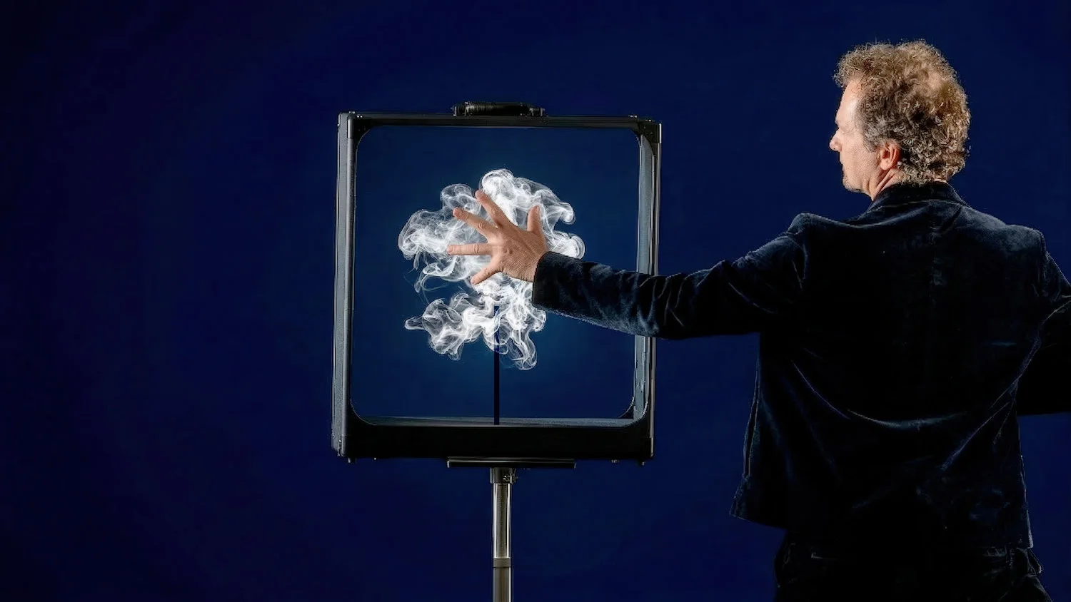 Le magicien, de dos, tend la main vers la valise ouverte posée sur pied : une volute de fumée blanche flotte à l&rsquo;intérieur du cadre.