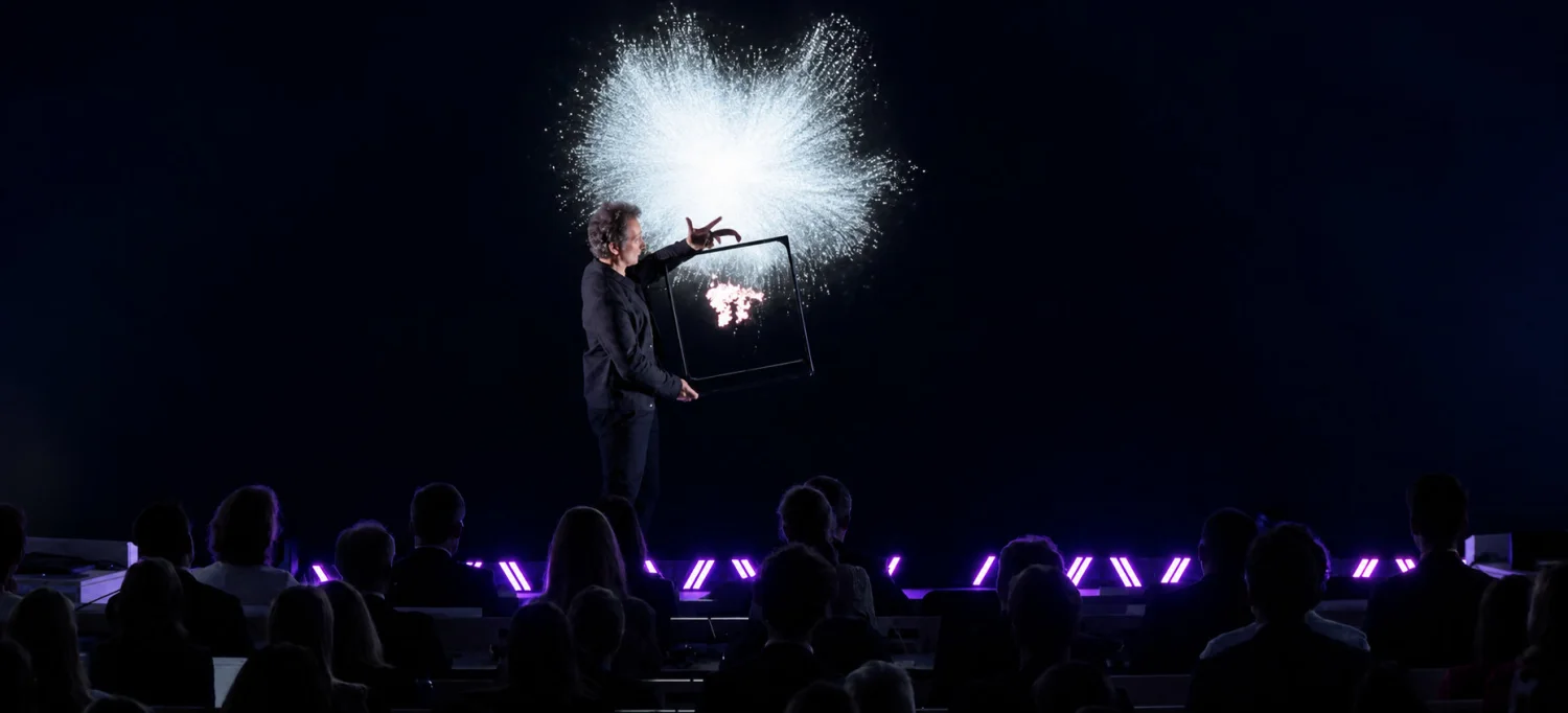 On stage, facing the seated audience, Romain Lalire holds the suitcase frame out at arm’s length: a burst of white particles explodes above it, carrying the hologram beyond the frame.