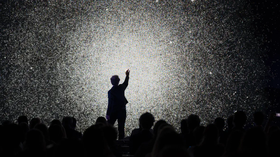 Un performeur en silhouette, bras levé, devant un mur d'image scintillant synchronisé avec les hélices.
